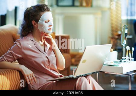 giovane donna in pigiama con maschera facciale in foglio bianco vicino al tavolo con articoli da toeletta e specchio utilizzando il computer portatile a casa moderna nella soleggiata giornata invernale. Foto Stock