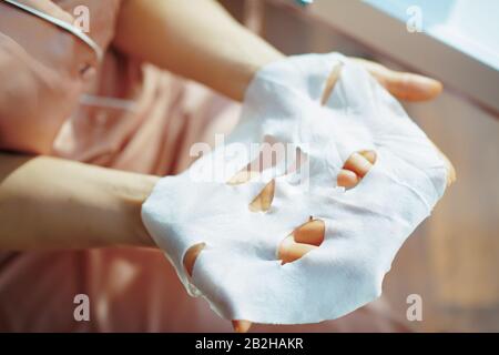 Closeup su elegante femmina in pigiami nella casa moderna nella soleggiata giornata invernale tenendo maschera facciale in foglio bianco. Foto Stock