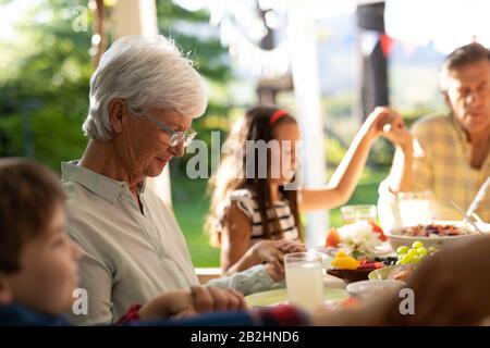 Vista laterale di una nonna e nonno caucasici, e loro nipote e figlia giovani seduti fuori ad un tavolo da pranzo fissato per un pasto con gli occhi Foto Stock