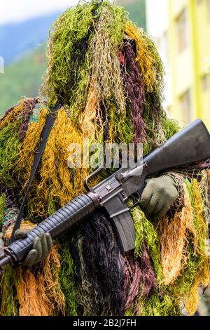 Un soldato che indossa una tuta ghillie completa in materiale fibroso verde, giallo, marrone e nero tiene un fucile nero con entrambe le mani. La tuta copre tutto il corpo e la testa, con solo un piccolo Foto Stock