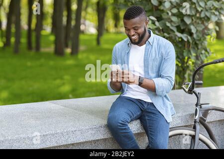 Gioioso ragazzo nero parcheggiava la bicicletta vicino al banco e utilizzando lo smartphone nel parco Foto Stock