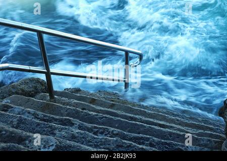 Un vortice di acqua di mare, schiume e lavaggi intorno ai gradini e corrimano di Giles Bagni, piscina di roccia oceanica Coogee Bay, Sydney, Australia Foto Stock