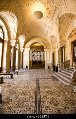 Un ampio corridoio interno ben illuminato caratterizzato da elementi architettonici classici. Lo spazio è definito da un alto soffitto ad arco decorato con modanature decorative e un pendente sferico centrale Foto Stock