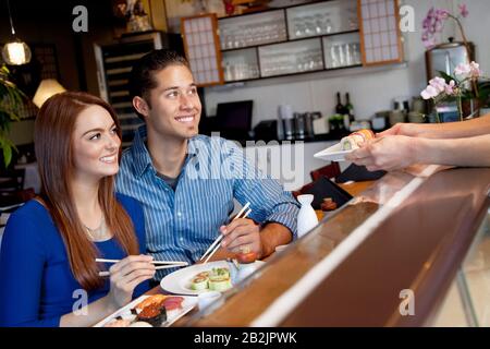Coppia giovane e felice che guarda lo chef che serve loro sushi nel ristorante Foto Stock