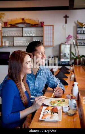 Coppia giovane e felice seduti al banco con sushi mentre si guarda via nel ristorante Foto Stock