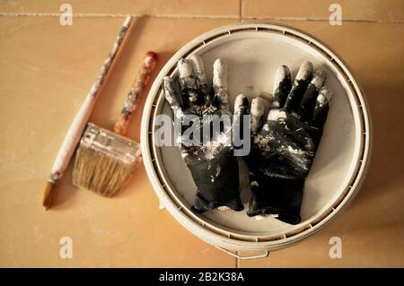 Verniciatura sul pavimento dopo l'uso. Dipingere guanti, barattolo e spazzole con vernice bianca in tutta la casa in costruzione. Foto Stock