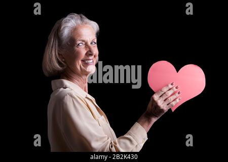 Ritratto di donna anziana che tiene carta rossa del cuore in studio Foto Stock