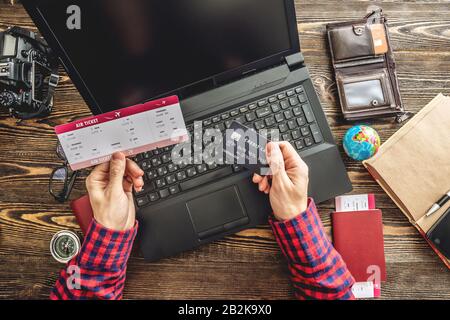 Prepararsi per un viaggio emozionante. L'uomo prenota i voli digitando su un computer portatile e pagando con una carta di credito su uno sfondo di legno. La vista dall'alto Foto Stock