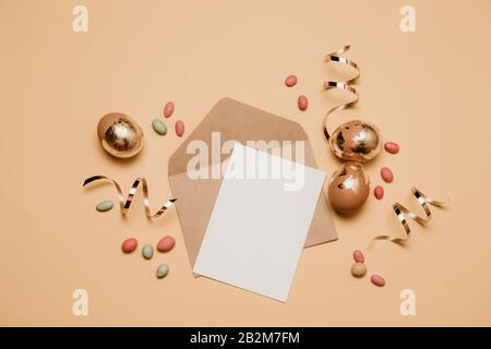 Busta bianca artigianale con foglio di carta da lettura, nastro oro glitter su sfondo beige. Uova d'oro di pasqua con caramelle di gelatina fagioli, vista dall'alto. Foto Stock