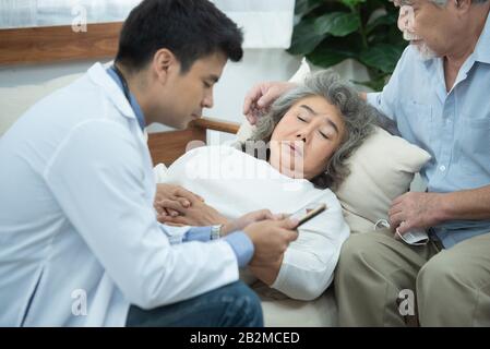 Anziano asiatico anziano paziente donna deposto sul divano in casa in attesa di risultato da giovane astuto medico caucasico smartphone Foto Stock
