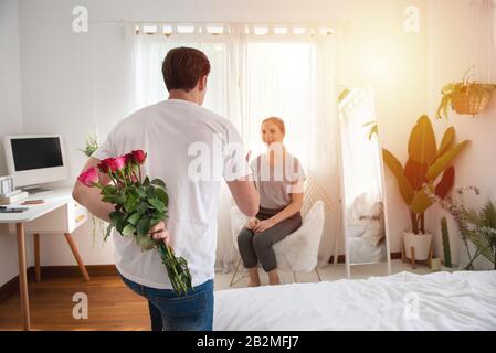 Momento inatteso nella vita quotidiana di routine! foto delle mani dell'uomo che si nascondono tenendo bouquet chic di rose rosse con dietro, donna felice è sfocata Foto Stock