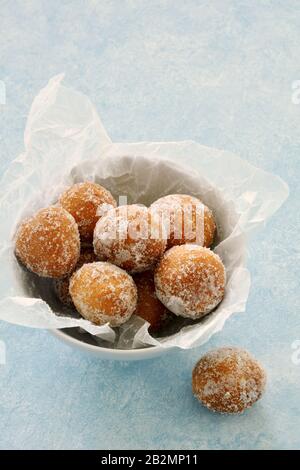 Buchi di ciambella di zucchero fresco in ciotola bianca con carta di cera sbriciolata su fondo azzurro in formato verticale. Foto Stock