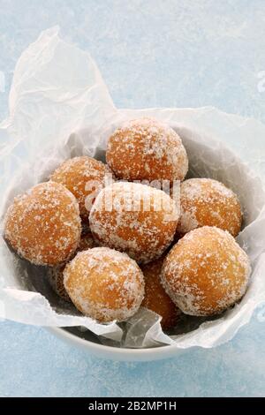 Buchi di ciambella di zucchero fresco in ciotola bianca con carta di cera sbriciolata su fondo azzurro in formato verticale. Foto Stock