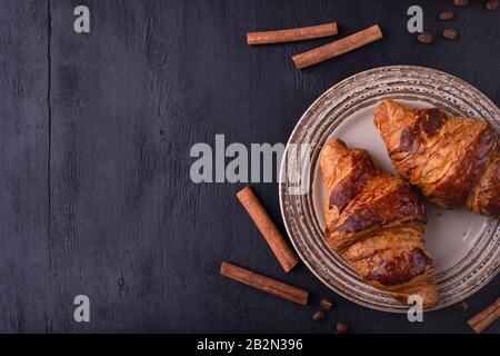 Cornetti su un piatto di ceramica, bastoncini di cannella e chicchi di caffè su un tavolo di legno nero. Vista dall'alto con spazio per il testo. Sfondo per ristorante, cucina Foto Stock