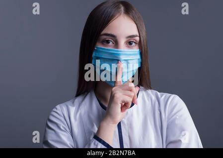 Ritratto di una giovane ragazza. Dottore in cappotto bianco e maschera medica. Guardando la macchina fotografica. Foto studio su sfondo grigio. Precauzioni wh Foto Stock