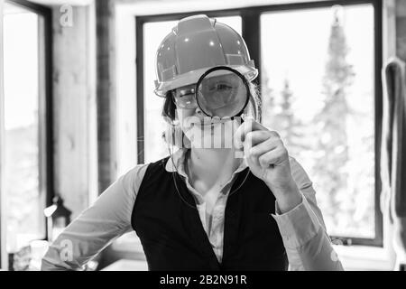 Occhio ingrandito di donna ispettore di costruzione che guarda attraverso lente d'ingrandimento che indossa il cappello duro durante l'ispezione di qualità dell'aria in bianco e nero Foto Stock