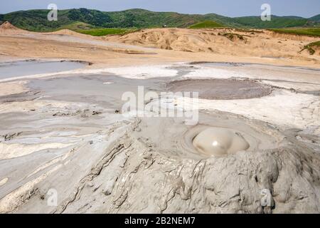Un'ampia vista di un'area geotermale con vulcani di fango attivi con gorgoglianti e viscosi piscine di fango. Il primo piano mostra una cupola di fango prominente con motivi vorticosi, circondata da fango incrinato e secco Foto Stock