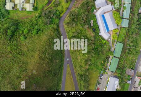 Una vista aerea dall'alto verso il basso mostra una stretta e tortuosa strada asfaltata che attraversa un lussureggiante paesaggio verde. La strada curva attraverso una fitta vegetazione e aree erbose, delimitate da gruppi di edifici con Foto Stock
