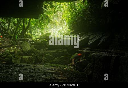 Una persona che indossa un casco rosso e abiti scuri sta navigando su terreni rocciosi ricoperti di muschio all'interno di un ingresso alla grotta. La scena è illuminata dalla luce naturale che filtra attraverso una fitta tettoia della giungla Foto Stock