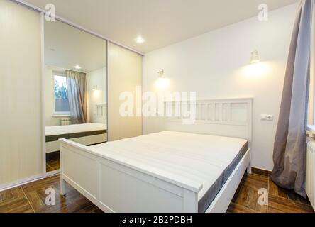 camera da letto in colori bianchi nella casa. Grande letto matrimoniale bianco, grande specchio sul guardaroba scorrevole, pavimento in parquet beige. Foto Stock