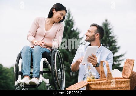 uomo allegro che usa un tablet digitale mentre riposa nel parco con la ragazza disabile Foto Stock