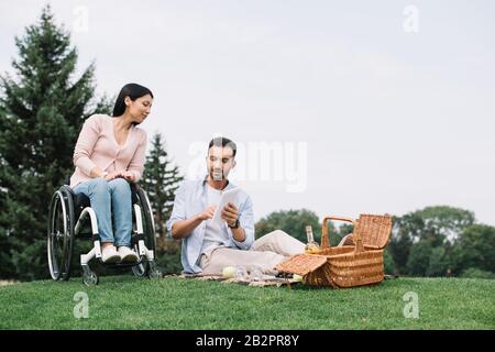 bel giovane uomo che usa un tablet digitale mentre riposa con la ragazza disabile nel parco Foto Stock