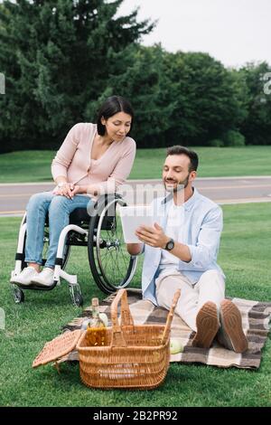 bel giovane che tiene un tablet digitale mentre si riposa con la ragazza disabile nel parco Foto Stock