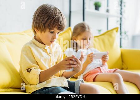 fuoco selettivo di ragazzo carino con sorella seduto sul divano e utilizzando gli smartphone a casa Foto Stock