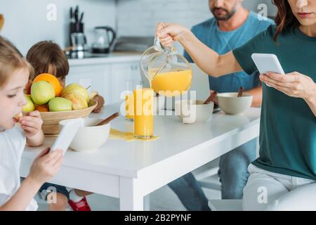 vista corta della donna con smartphone in vetro overfilling con succo d'arancia mentre si siede al tavolo da cucina vicino alla famiglia utilizzando smartpones Foto Stock