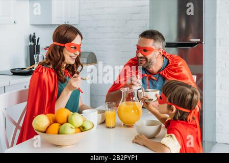 famiglia allegra in costumi di supereroi con colazione in cucina Foto Stock