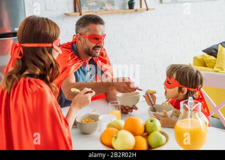 famiglia allegra in costumi supereroi con colazione in cucina insieme Foto Stock