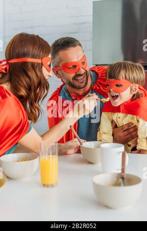 famiglia allegra in costumi di supereroi a colazione, mentre madre nutriva figlio con fiocchi Foto Stock