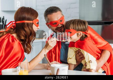 madre nutrendo il figlio con i fiocchi, mentre la famiglia in costumi di supereroi che hanno colazione Foto Stock