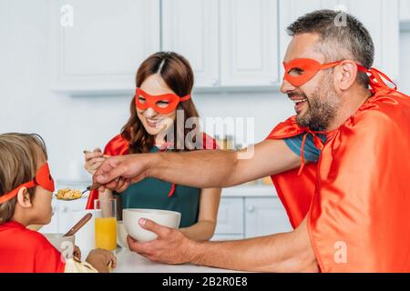 famiglia in costumi supereroi che hanno colazione mentre padre che dà da mangiare a figlio con fiocchi Foto Stock