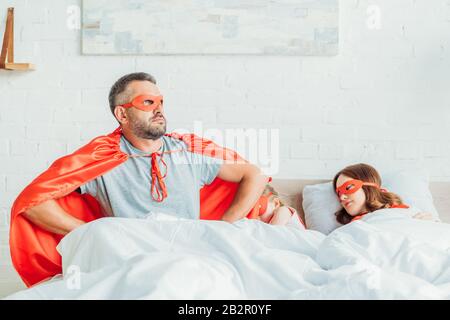 padre serio in costume di supereroe seduto a letto vicino alla famiglia dormiente, tenendo le mani sui fianchi e guardando lontano Foto Stock
