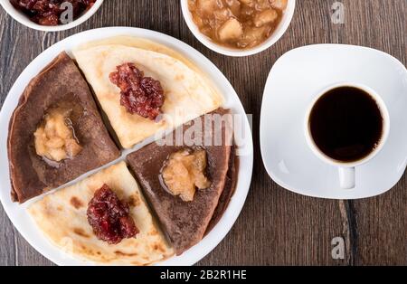 Disposizione piatta. I pancake sono semplici e con cacao. Frittelle su un piatto bianco, marmellata di mele di diverse varietà e caffè su uno sfondo di legno. Traditio Foto Stock