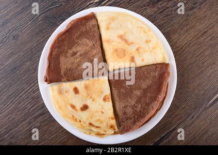 Disposizione piatta. I pancake sono semplici e con cacao. Pancake su un piatto bianco su uno sfondo di legno. Colazione tradizionale russa. Foto Stock