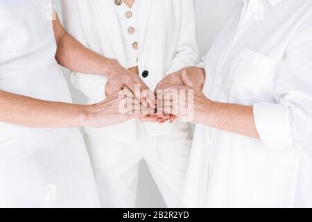 vista ritagliata di tre generazioni di donne in abiti bianchi che tengono le mani isolate sul grigio Foto Stock