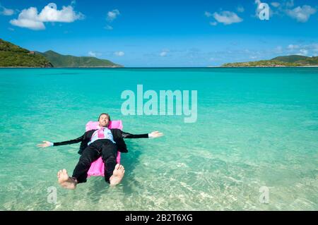 Uomo d'affari a piedi nudi che galleggia su una zattera rosa brillante in acque blu tropicali Foto Stock