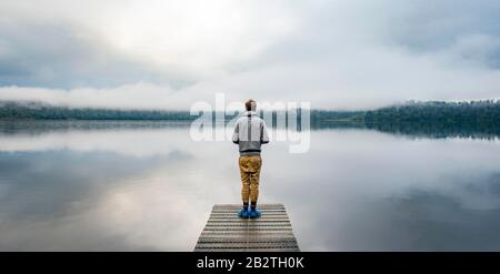 Giovane uomo in piedi su un molo che si affaccia su un lago, nebbiosa atmosfera, Lago Mapourika, Costa Occidentale, Isola del Sud, Nuova Zelanda Foto Stock