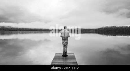 Giovane uomo in piedi su un molo che si affaccia su un lago, nebbiosa atmosfera, Lago Mapourika, Costa Occidentale, Isola del Sud, Nuova Zelanda Foto Stock