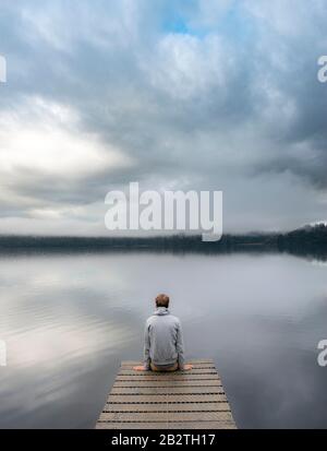 Giovane uomo seduto su un molo che si affaccia su un lago, nebbiosa atmosfera, Lago Mapourika, Costa Occidentale, Isola del Sud, Nuova Zelanda Foto Stock