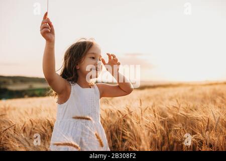 Sorridente ragazza caucasica vestita di abito bianco sta camminando in un campo di grano con un sorriso allegro sui salti Foto Stock