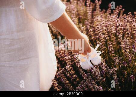 Sorreggia la madre che tiene un paio di scarpe del bambino e che cammina in un campo di lavanda mentre indossa un vestito bianco piacevole Foto Stock