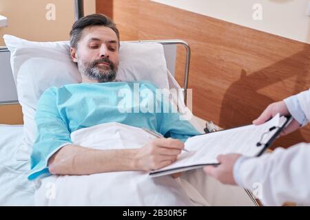 Un uomo maturo e serio nel letto dell'ospedale che firma l'accordo medico prima dell'operazione chirurgica Foto Stock