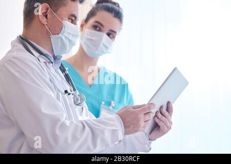 Medico in maschera e camice da laboratorio che usa la compressa digitale mentre discute l'epidemia di influenza con il collega in clinica Foto Stock