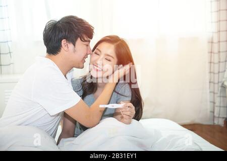 Allegro e felice giovane asiatico uomo e donna con test di gravidanza positivo tenendo in mano mentre seduto a letto in camera da letto a casa. Foto Stock