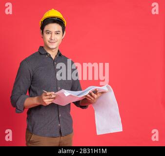 Giovane uomo asiatico bello in abito casual con cappello giallo mano tenendo carta piano in architetto e ingegnere lavoratore isolato in rosso Foto Stock