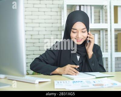 Occupato giovane asiatico musulmano business donna parlare sul cellulare e utilizzando il computer portatile in ufficio moderno. Foto Stock