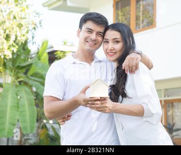 Felice giovane caucasico uomo abbraccio donna asiatica con amore mano tenendo la forma della scatola casa mostrando all'aperto con nuova casa in background e guardare la macchina fotografica. Foto Stock